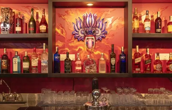 A bar scene with an array of liquor bottles on shelves, in front of a vibrant mural of a fiery, stylized creature. Below, a neat arrangement of glasses and a beer tap await use.