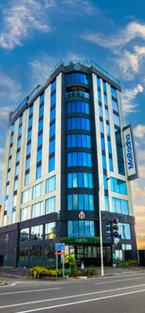 A vertical image of the Maradha Hotel building, showcasing its blue and white facade with large windows, set against a backdrop of scattered clouds in the sky.