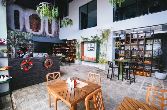 A rustic outdoor café area featuring wooden tables and chairs, a bar with floral decorations, and shelves stocked with various items. The space is accented with green plants and has an urban yet cozy feel.