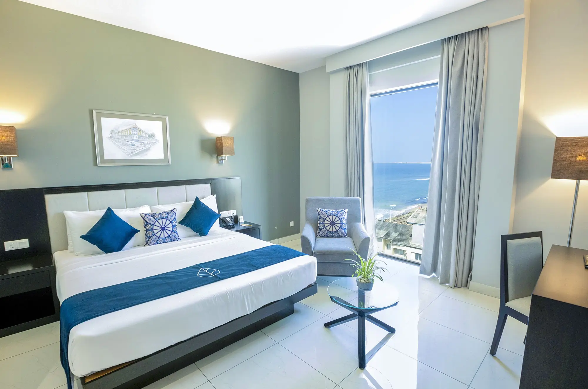 A deluxe queen room at Hotel Maradha, tastefully decorated with a queen-sized bed with white bedding and blue accents, matching accent pillows, a comfortable grey armchair, elegant lighting, and a window revealing a scenic view of the sea and coastal area below.