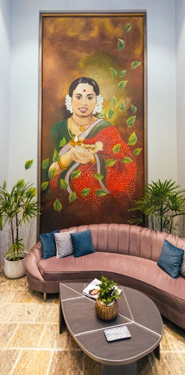 An elegant hotel lobby area with a curved pink sofa, a modern gray coffee table, and lush green plants. A large, traditional painting of a woman hangs prominently on the wall above the sofa.