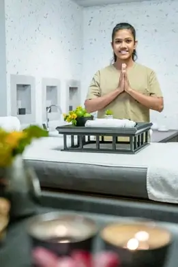 A welcoming spa therapist with hands clasped in a greeting gesture stands behind a massage table, with a focus on tranquility and relaxation indicated by the blurred foreground of lit candles.