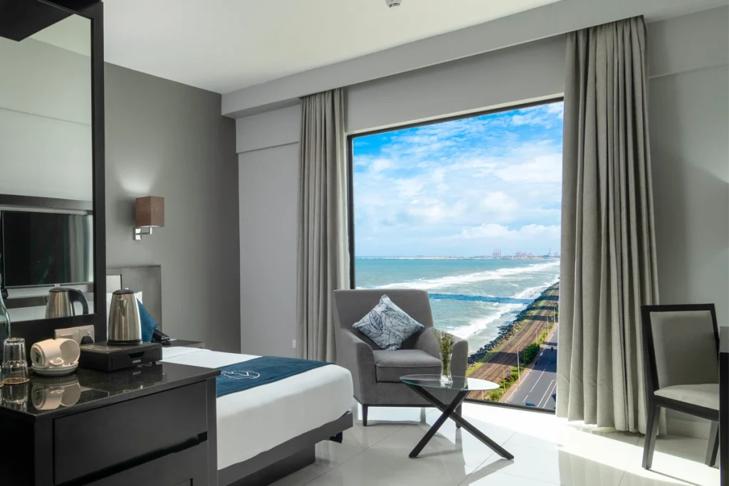 A deluxe king room at Hotel Maradha, featuring a large bed with white and navy blue bedding, a comfortable grey armchair, modern amenities, and a large picture window with a stunning view of the coastal shoreline and blue sea.