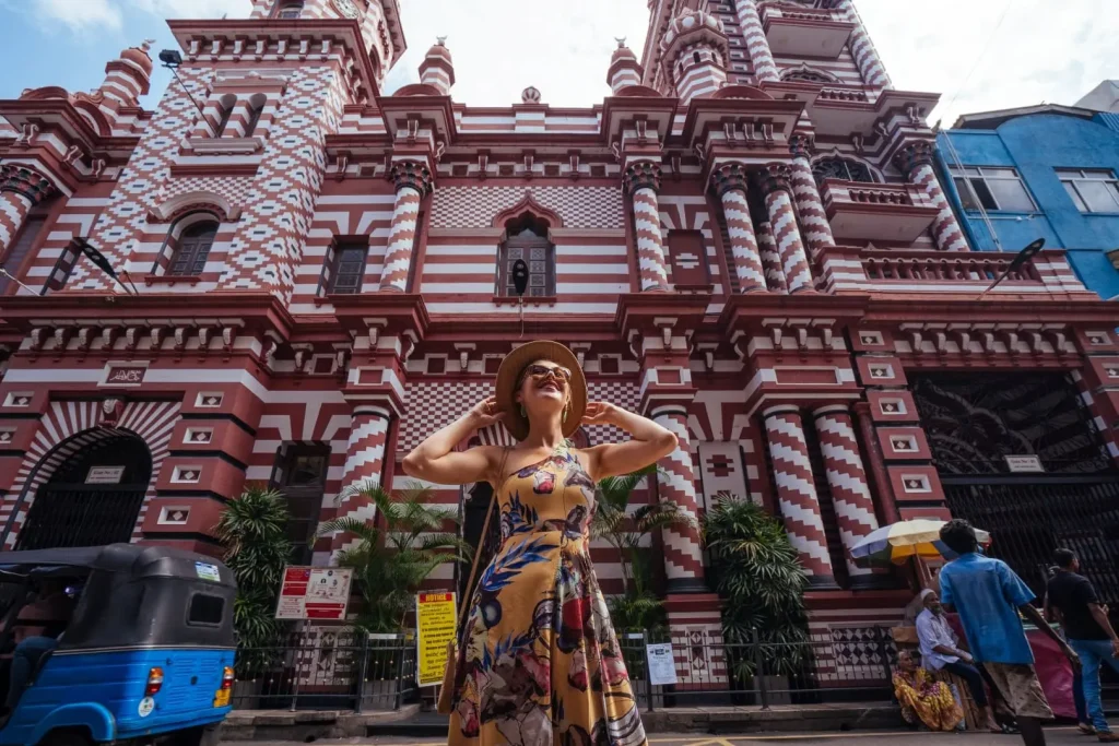 A tourist in a summer dress and hat stands admiringly in front of the striking red and white patterned facade of the Jami Ul-Alfar Mosque in Colombo, under a bright blue sky.