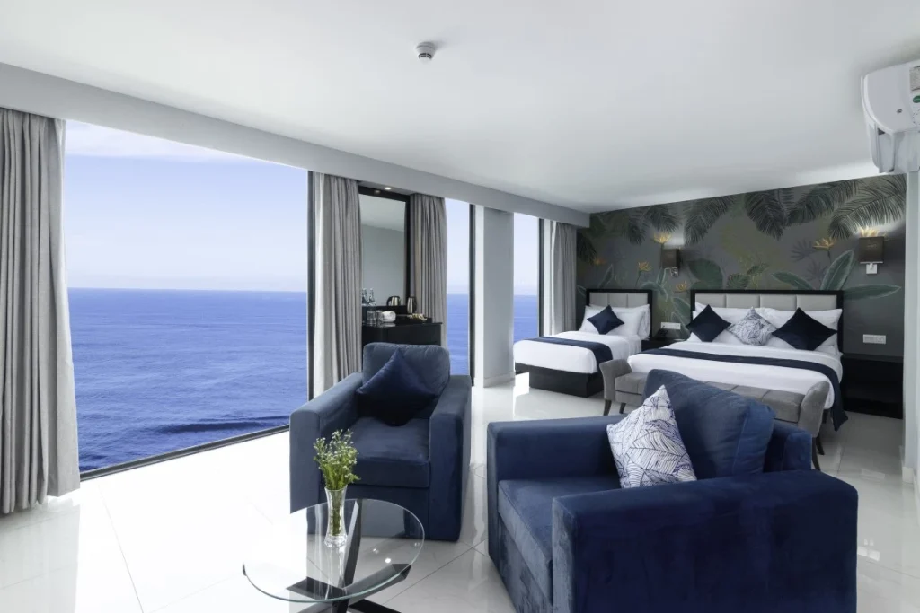 The Signature Family Sea Front room at Hotel Maradha featuring two double beds with white and navy blue bedding against a tropical-themed accent wall, comfortable blue armchairs, a glass coffee table with a bouquet, and expansive windows offering a majestic view of the sea.