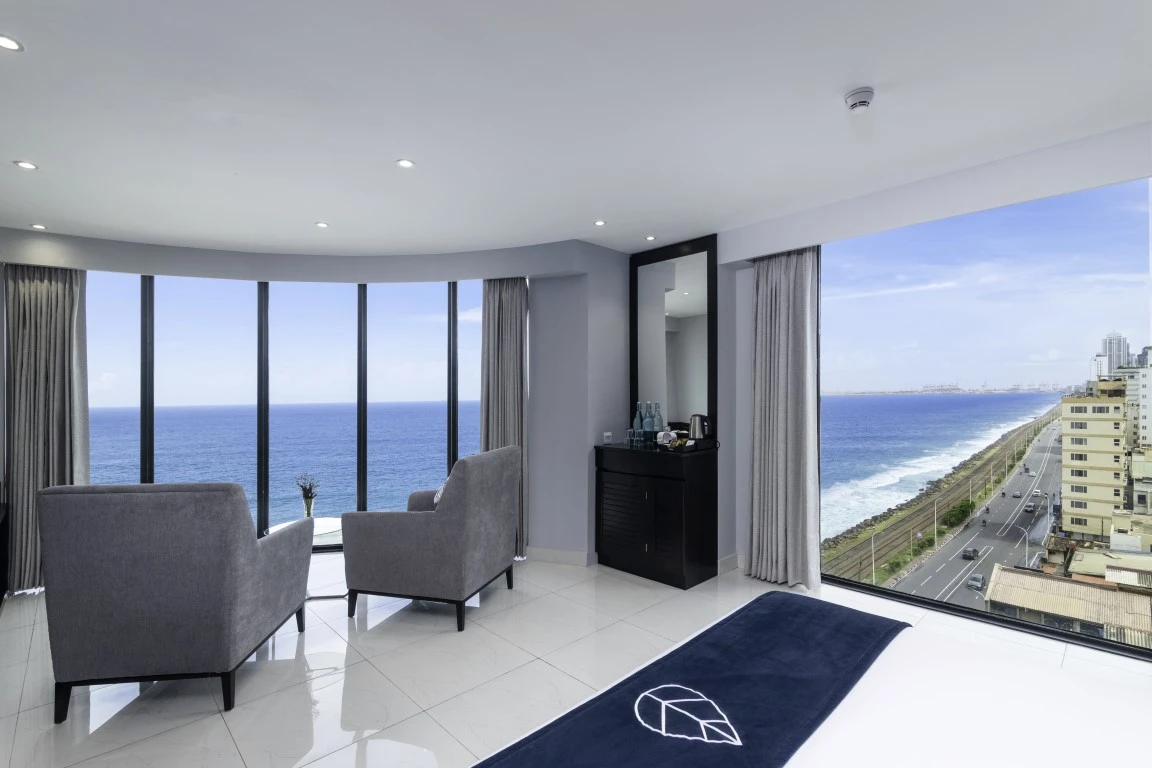 The Signature Panorama Sea Front room at Hotel Maradha offers a stunning 180-degree view of the ocean through wall-to-wall windows, with two plush armchairs, a sleek service console, and the room's décor featuring a calming palette of grays and whites, complemented by a striking navy blue bed runner with the hotel's logo.