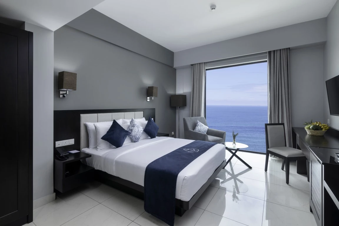 Elegant premier two-bedroom seafront suite in Hotel Maradha with a plush king-size bed adorned with navy blue and white linens, accent pillows with geometric patterns, a cozy sitting area, contemporary furnishings, and a panoramic window offering a breathtaking view of the sea.