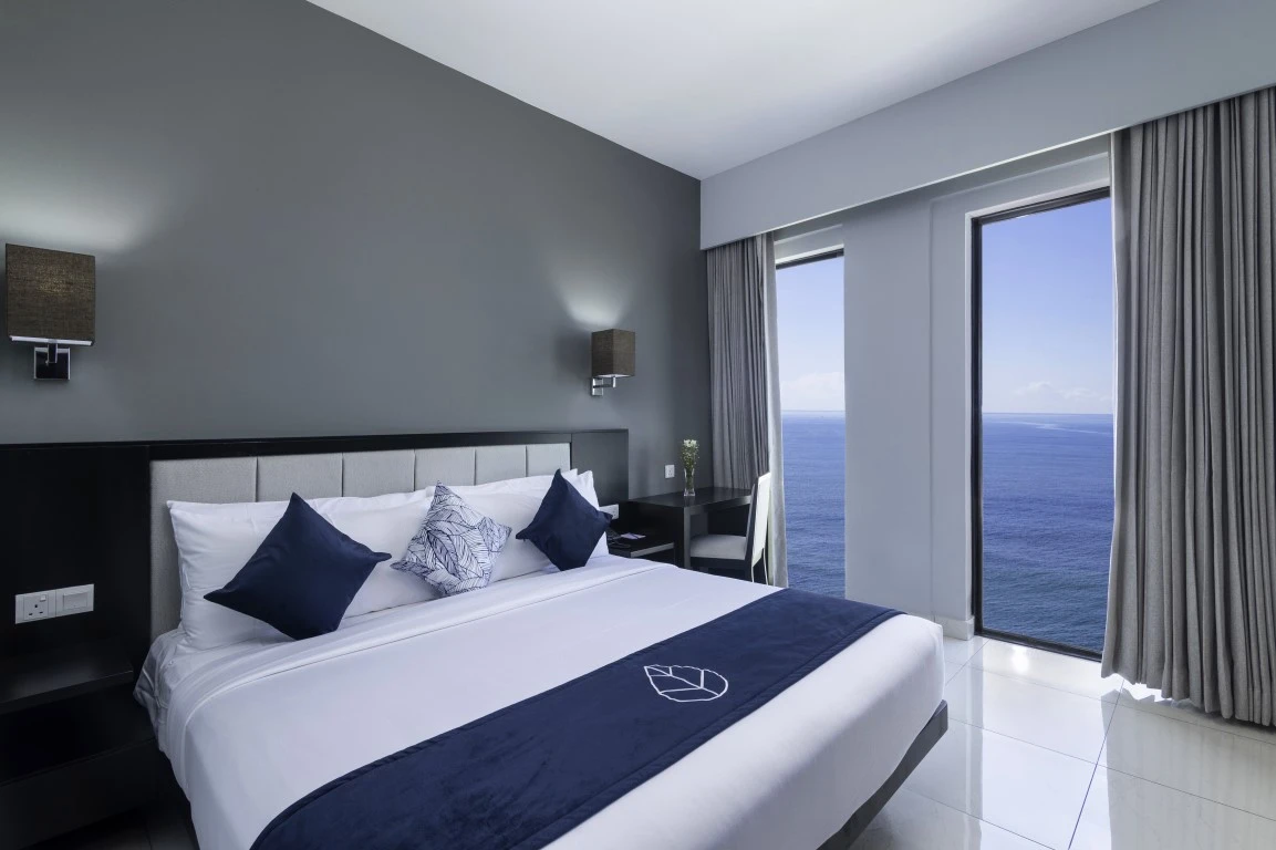 A sophisticated premier one-bedroom seafront suite in Hotel Maradha, displaying a luxurious king-size bed with crisp white sheets, navy blue accents, patterned throw pillows, sleek dark-toned furniture, and floor-to-ceiling windows providing a stunning view of the endless ocean.